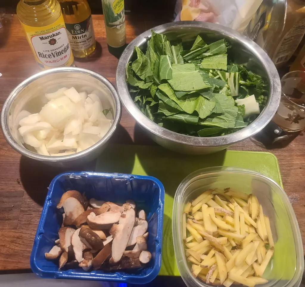 Prepped ingredients on the countertop in various metal and plastic containers. From upper left to lower right: white Vidalia onion, chopped bok choy, sliced shiitake mushroom, fresh julienned ginger sticks.