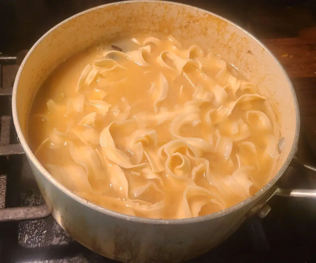 A saucepan full of miso broth boils while udon are steeping in it. The noodle are not yet fully cooked, and there is plenty of liquid. A star anise is visible in the broth.
