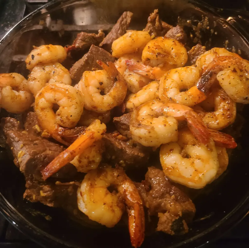 Cooked shrimp are added to the beef. No additional garnish has been added yet and they are lying atop the steak.