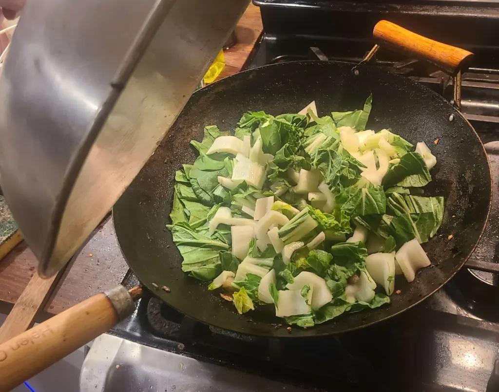 A wok's lid has been raised to show chopped bok choy, yet to be cooked, resting in some aji mirin over onions and other veggies.