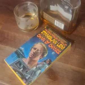 The paperback version of Chain of Chance is shown on a counter with a glass and a bottle of Woodford Reserve Select bourbon.