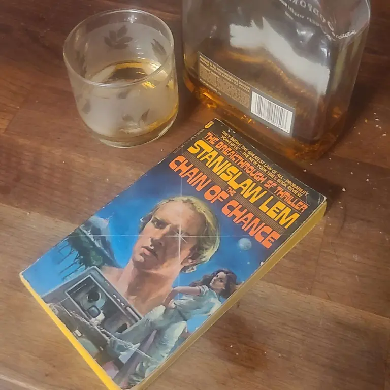 The paperback version of Chain of Chance is shown on a counter with a glass and a bottle of Woodford Reserve Select bourbon.